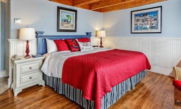 A king bed with a red quilt and nautical themed pillows sits at the center of a hotel room. Wood beams frame the ceiling and there are beautiful hardwood floors. A pair of white nightstands flanks the bed.