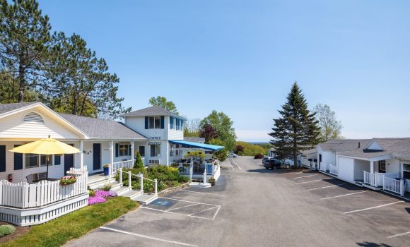 The outside view of a midcoast Maine area hotel. The lobby has a covered porch and rooms open directly into the parking lot. The hotel is white with navy siding.