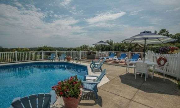 A hotel pool on a sunny summer day. Deck chairs and umbrellas are lined up around the patio. A pot of flowers is in the foreground.